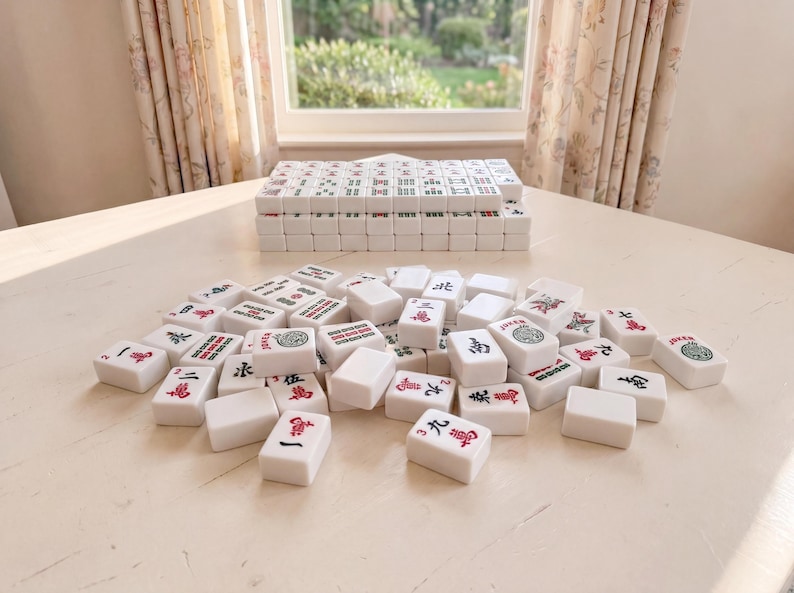 May include: A complete Mahjong set with white tiles featuring red and green symbols and characters. The tiles are scattered on a light-colored surface, with a stack of tiles in the background. The game is set up on a table near a window.