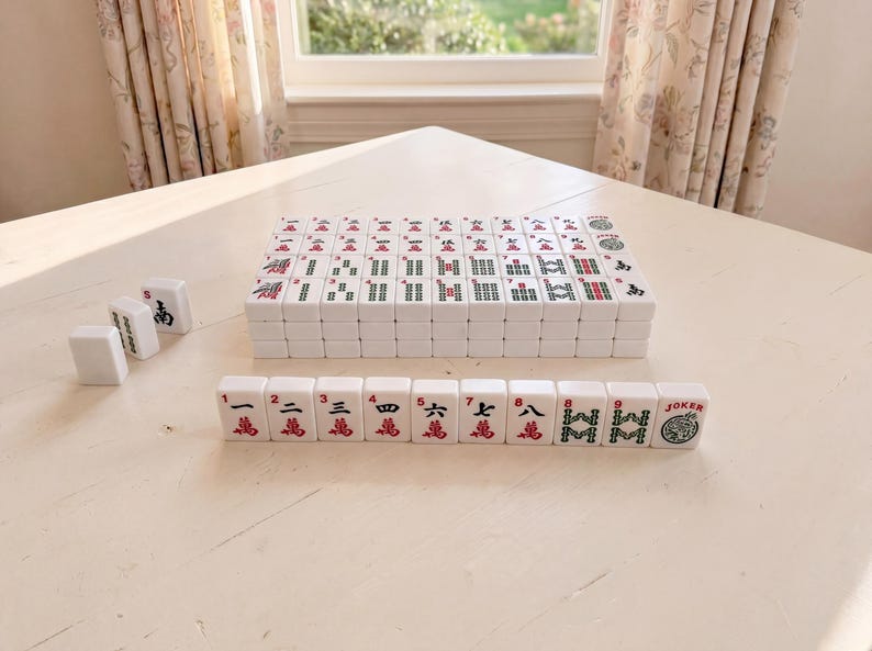 May include: A set of white Mahjong tiles with red and green markings, arranged on a white table. The tiles are stacked and laid out, showcasing the game's components. The scene is lit by natural light, suggesting an indoor setting.