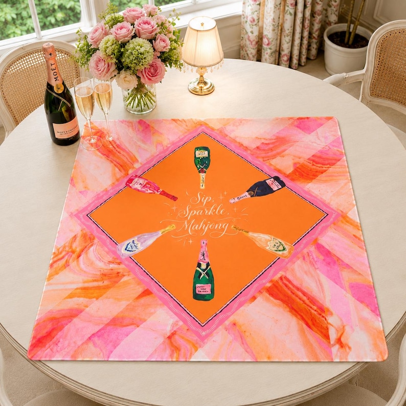 May include: A square, orange and pink patterned table mat with a diamond-shaped design featuring champagne bottles. The mat has the words "Sip, Sparkle, Mahjong" in cursive. A bottle of champagne and two glasses are on the table.
