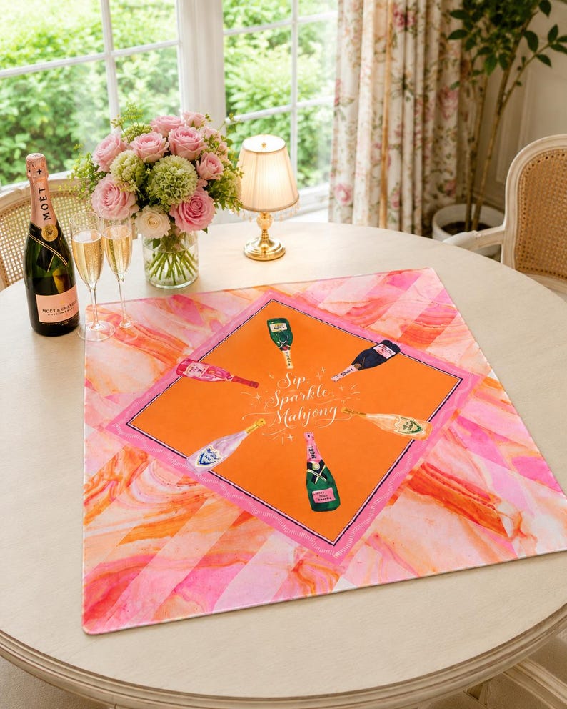 May include: A round table setting with a pink and orange patterned table runner. The runner features champagne bottles and the text "Sip, Sparkle, Mahjong." A bottle of Mo&euml;t champagne, two glasses, and a bouquet of pink roses are also on the table.