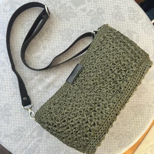 May include: Olive green crochet crossbody bag with a black leather strap and accents. The bag features a detailed starburst pattern and a rectangular shape. The strap is adjustable and attached with silver-tone hardware.