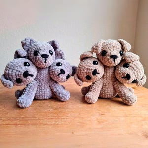 Op de afbeelding: Twee gehaakte Cerberus hondenknuffels. Eén is lavendelkleurig, de andere is bruin. Elk heeft drie hoofden met zwarte ogen en neuzen. De knuffels staan op een houten oppervlak.