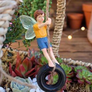 May include: A miniature fairy figurine with translucent wings, wearing a yellow shirt and blue shorts, swings on a tire swing. The fairy is suspended from a rope, with a backdrop of succulents and a woven basket.