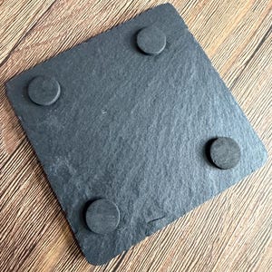 Peut inclure: Un dessous de verre carr&eacute; en ardoise gris fonc&eacute; avec une texture de pierre naturelle rugueuse. Le dessous pr&eacute;sente quatre petits patins en mousse noire circulaires plac&eacute;s pr&egrave;s de chaque coin pour assurer la stabilit&eacute;.