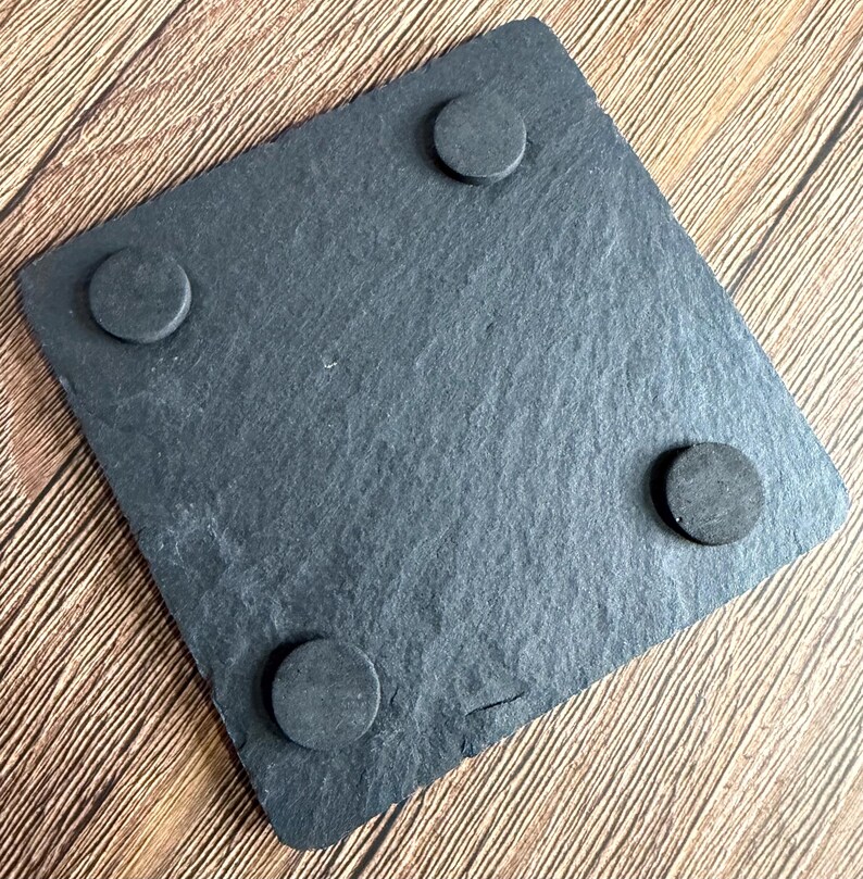 Peut inclure: Un dessous de verre carr&eacute; en ardoise gris fonc&eacute; avec une texture de pierre naturelle rugueuse. Le dessous pr&eacute;sente quatre petits patins en mousse noire circulaires plac&eacute;s pr&egrave;s de chaque coin pour assurer la stabilit&eacute;.