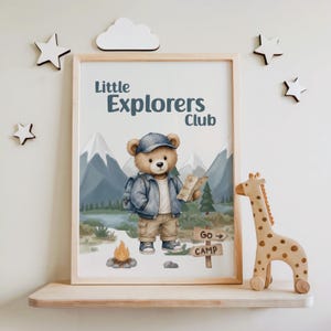 Peut inclure: Une affiche encadrée représentant un ours brun portant une casquette, une veste et un sac à dos bleus, tenant une carte. Le texte indique Little Explorers Club et Go Camp. Le décor montre des montagnes, des arbres et un feu de camp.