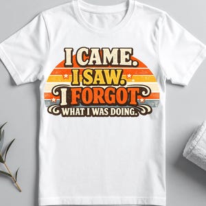 Peut inclure: Un t-shirt blanc à manches courtes avec un motif graphique sur fond de coucher de soleil rétro aux tons orange, jaune et bleu. Le texte indique I CAME. I SAW. I FORGOT WHAT I WAS DOING. Les lettres sont marron foncé avec un aspect vieilli.