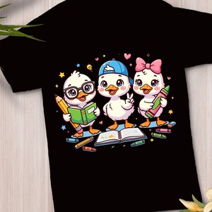Peut inclure: Un t-shirt noir avec un motif représentant trois canards blancs de dessin animé. Un canard porte des lunettes et tient un livre vert et un crayon jaune, un autre porte une casquette bleue, et le troisième porte un nœud rose en tenant un crayon rose. Des étoiles et des cœurs entourent les personnages, avec un cahier ouvert et des crayons au sol.