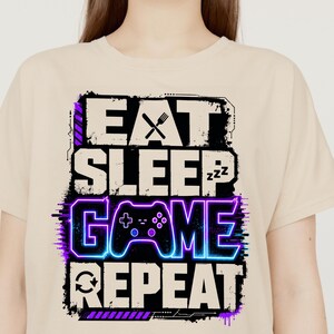Peut inclure: Un t-shirt beige avec une impression graphique portant le texte EAT SLEEP GAME REPEAT. Le motif comprend une icône de fourchette dans la lettre A, des symboles ZZZ dans la lettre P, une manette de jeu bleue et violette et une flèche circulaire.