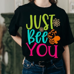 Peut inclure: Un t-shirt noir à manches courtes avec le texte "JUST Bee YOU" écrit en vert lime, turquoise et rose vif. Le motif comprend une petite abeille orange dessinée et deux icônes de fleurs beiges placées à côté du mot "JUST".