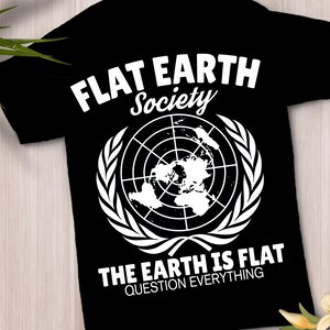 Peut inclure: Un t-shirt noir avec un graphisme blanc représentant une carte de la terre plate dans une grille circulaire, entourée de feuilles de laurier. Le texte indique FLAT EARTH Society, THE EARTH IS FLAT et QUESTION EVERYTHING en blanc.