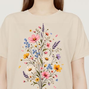 Peut inclure: Un t-shirt beige à manches courtes orné d'un imprimé central représentant un bouquet de fleurs sauvages colorées. L'illustration comprend des cosmos roses, des marguerites blanches, de la lavande violette, des myosotis bleus et des fleurs jaunes.