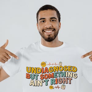 Peut inclure: Un t-shirt blanc avec un motif portant le texte "UNDIAGNOSED BUT SOMETHING AIN'T RIGHT" dans une police rétro multicolore. Le design comprend deux visages emoji jaunes portant des lunettes, deux lignes EKG cardiaques rouges et deux presse-papiers médicaux.