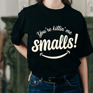 Peut inclure: Un t-shirt noir à manches courtes avec le texte couleur crème "You're killin' me smalls!" imprimé sur la poitrine dans une police stylisée, accompagné d'un graphique en ligne courbe sous le texte qui ressemble à un sourire simple.