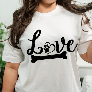 May include: A white short-sleeved t-shirt featuring a black graphic print on the chest. The design displays the word Love in a cursive script, where the letter o is replaced by a heart shape containing a paw print, positioned above a dog bone.