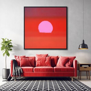 Pode incluir: Uma peça de arte de parede quadrada com um fundo vermelho sólido e uma grande forma circular rosa claro centrada na parte superior. A obra de arte está inserida numa moldura preta fina e é exibida acima de um sofá vermelho com almofadas.