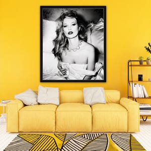 May include: A black and white framed photograph featuring a portrait of a person with wavy hair wearing a necklace and draped in white fabric, positioned against a background of pillows. The artwork is displayed in a thin black frame.