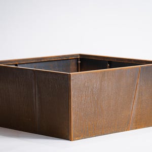 Puede incluir: Jardinera cuadrada de metal con acabado marrón oxidado. Los lados se inclinan ligeramente hacia afuera y el interior es oscuro. Aspecto rústico, adecuado para uso en exteriores o interiores. Dimensiones: aproximadamente 60 cm x 60 cm.