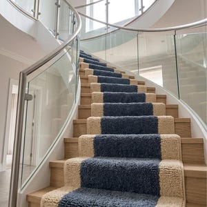 May include: A curved staircase featuring light oak wooden steps and a central runner made of textured, deep blue and beige carpet. The staircase is enclosed by a curved glass railing with a silver metal handrail and a large arched window above.
