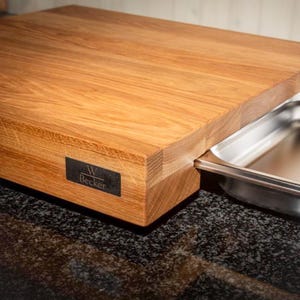 Puede incluir: Una tabla de cortar de madera gruesa y rectangular con un acabado de grano de madera marrón claro natural. Una placa de metal rectangular negra con el texto blanco W Becker está unida al lateral. Una bandeja de acero inoxidable está insertada.