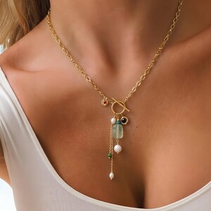 May include: A gold-toned chain necklace featuring a toggle clasp closure. The necklace is adorned with a central pendant arrangement consisting of a pale green rectangular stone, white pearls, a small black circular charm, and a tiny red accent.