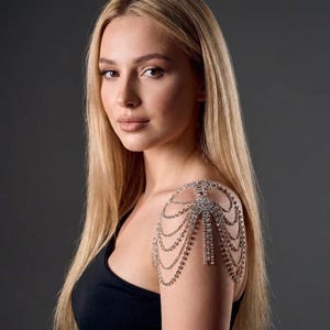 May include: A silver rhinestone shoulder piece with multiple cascading strands. The shoulder jewellery is attached to a central design and drapes over the shoulder and arm. The model is wearing a black vest top.
