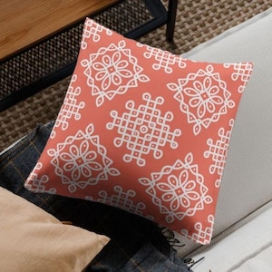Op de afbeelding: Vierkant koraalkleurig sierkussen met een wit geometrisch patroon. Het patroon heeft bloem- en vierkante vormen. Het kussen ligt op een lichtgrijze bank. Het kussen is een decoratief item.
