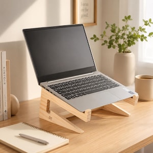 May include: A wooden laptop stand supporting a silver laptop. The stand is angled for ergonomic use. The desk setup includes a notebook, pen, and a framed print with the words "Focus on the good."