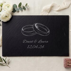 Puede incluir: Un recuerdo de boda de pizarra negra con anillos grabados, los nombres "David & Laura" y la fecha "12.04.24". Flores blancas y una cinta están dispuestas alrededor de la pizarra. También hay un pequeño joyero rosa.