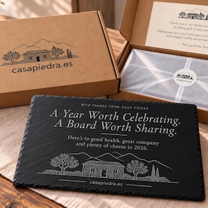 Puede incluir: Una tabla de servir rectangular de pizarra negra grabada con el texto A Year Worth Celebrating. A Board Worth Sharing. Here's to good health, great company and plenty of cheese in 2026. Se encuentra frente a una caja de regalo de cartón.