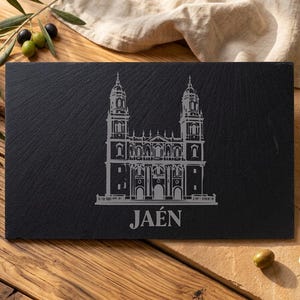 Puede incluir: Una tabla de servir rectangular de pizarra negra con un grabado detallado de una catedral y la palabra "JAÉN". La tabla está sobre una superficie de madera, con aceitunas y aceite de oliva cerca.
