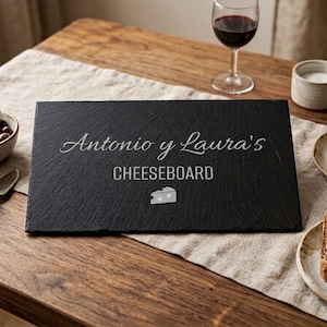 Puede incluir: Una tabla de queso rectangular de pizarra negra con el texto "Antonio y Laura's CHEESEBOARD" grabado en blanco. Sobre una mesa de madera, con una copa de vino tinto, uvas y pan.
