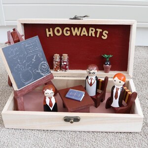 Può includere: Scatola di legno con interno rosso e scritta "HOGWARTS" in lettere di legno. All'interno ci sono figurine in miniatura, una lavagna su un cavalletto, un tavolo, sedie e piccoli barattoli di vetro. È visibile anche una pianta in miniatura.