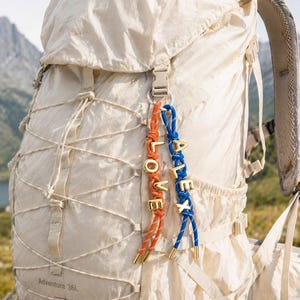 May include: A cream-colored backpack with a mountain backdrop. Two keychains hang from the backpack. One keychain is orange and gold with the word "LOVE". The other is blue and gold with the name "ALEX".