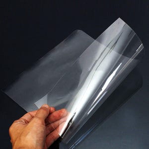 May include: A hand holding a clear, transparent sheet of plastic. The plastic sheet is slightly curved, reflecting light and creating highlights. The background is a dark, solid color.