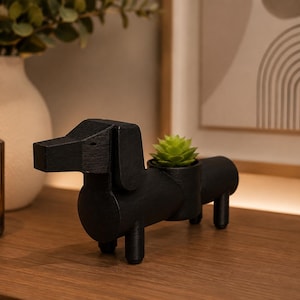 Könnte beinhalten: Ein schwarzer Dackel-Blumentopf aus Holz mit einer kleinen grünen Sukkulente. Der Hunde-Blumentopf hat ein minimalistisches Design und steht auf einer Holzoberfläche. Eine Kerze in einem Glashalter und eine weiße Vase befinden sich im Hintergrund.