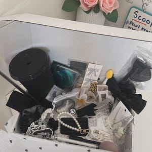 May include: A white box filled with various accessories, including black hair bows, scrunchies, and a black tumbler. Other items include a pearl bracelet with a smiley face charm, hair clips, and a makeup sponge. A vase of pink roses sits nearby.