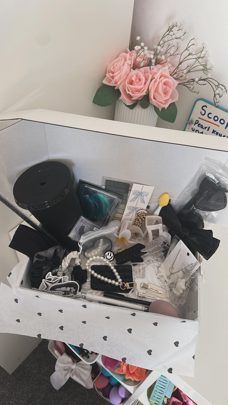 May include: A white box filled with various accessories, including black hair bows, scrunchies, and a black tumbler. Other items include a pearl bracelet with a smiley face charm, hair clips, and a makeup sponge. A vase of pink roses sits nearby.