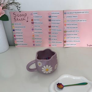May include: A lilac daisy-themed mug filled with colorful beads sits on a white surface, next to a white plate with a spoon. A sign lists various items, including necklaces and hair clips. A vase of pink roses is in the background.