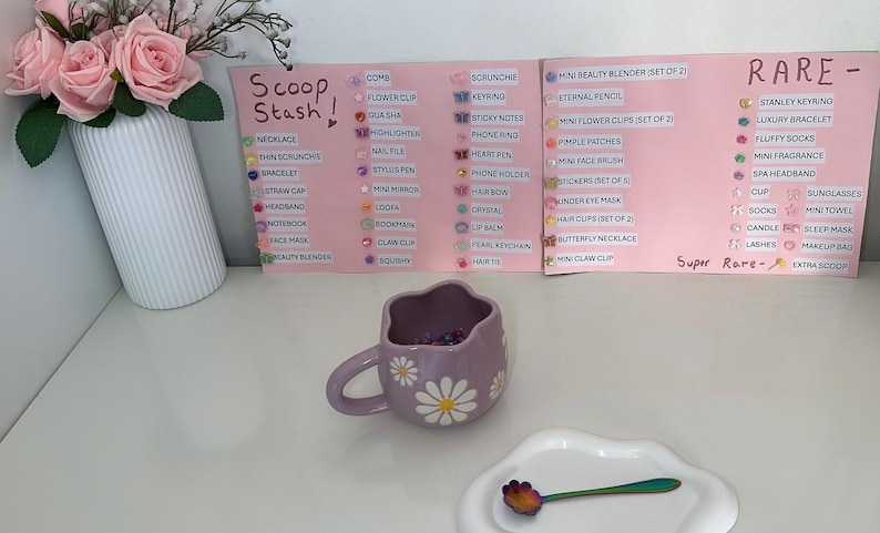 May include: A lilac daisy-themed mug filled with colorful beads sits on a white surface, next to a white plate with a spoon. A sign lists various items, including necklaces and hair clips. A vase of pink roses is in the background.
