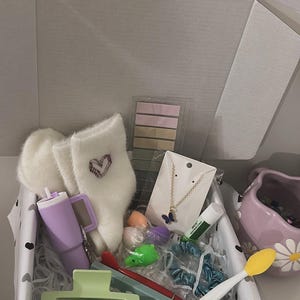 May include: An open gift box filled with various items. Includes fluffy white socks with a heart design, a purple tumbler, a butterfly necklace, hair accessories, and other small items in pastel colors. The box is lined with black and white polka dot paper.