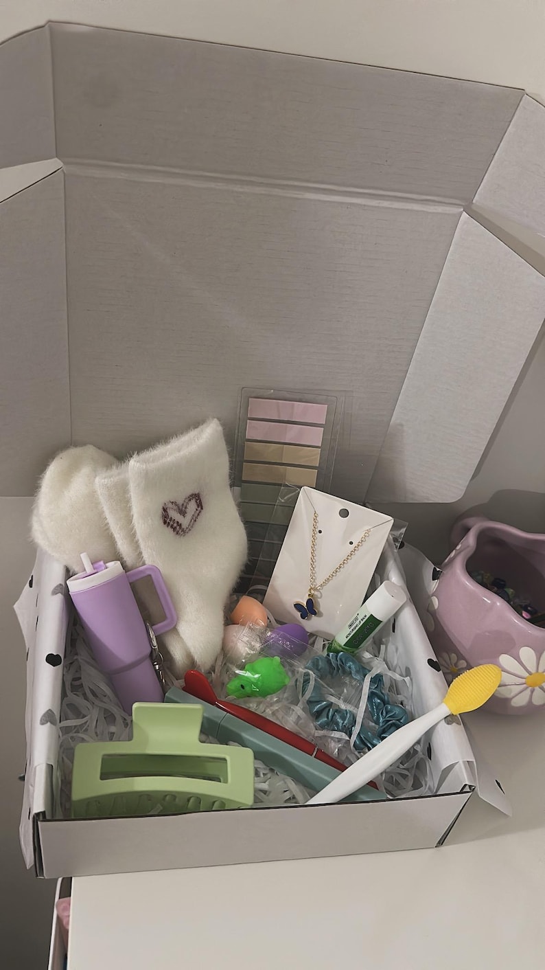 May include: An open gift box filled with various items. Includes fluffy white socks with a heart design, a purple tumbler, a butterfly necklace, hair accessories, and other small items in pastel colors. The box is lined with black and white polka dot paper.