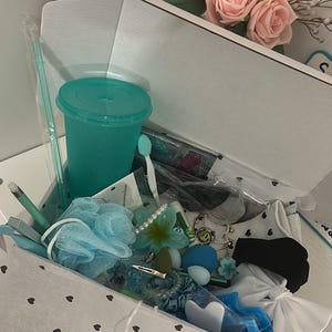 May include: A gift box filled with various teal and blue items, including a tumbler, bath sponge, hair accessories, and a toothbrush. A bouquet of pink roses sits nearby. The box is lined with white paper decorated with black hearts.