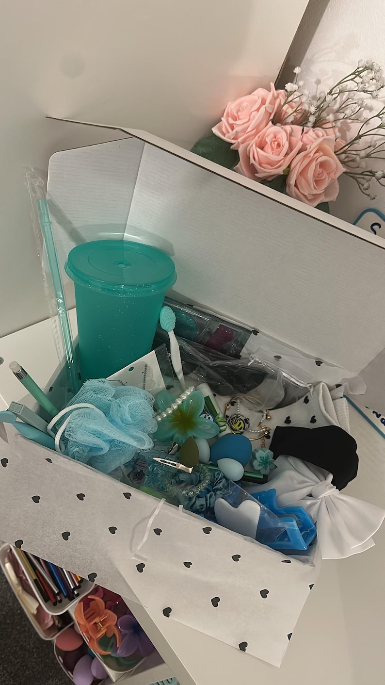 May include: A gift box filled with various teal and blue items, including a tumbler, bath sponge, hair accessories, and a toothbrush. A bouquet of pink roses sits nearby. The box is lined with white paper decorated with black hearts.