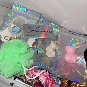 May include: A gift box filled with various items, including a pearl bracelet, a green loofah, scrunchies, and small bear-shaped soaps. A card with the words "Thank you" is also visible.