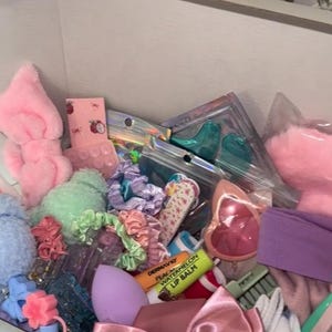May include: A box filled with various beauty accessories in pastel colors. Items include pink headbands, scrunchies, hair ties, a makeup sponge, lip balm, and a large pink satin bow. A bouquet of pink roses is visible in the background.