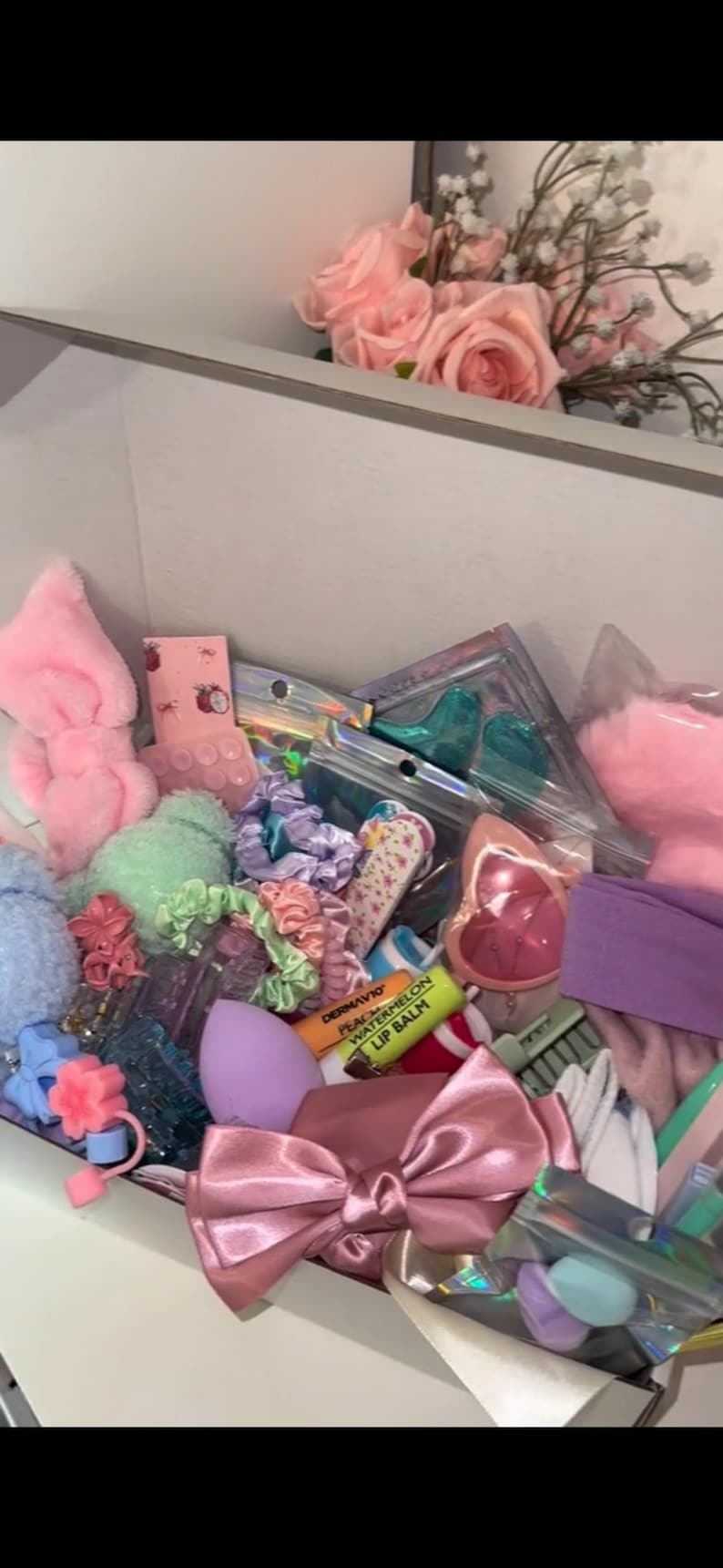 May include: A box filled with various beauty accessories in pastel colors. Items include pink headbands, scrunchies, hair ties, a makeup sponge, lip balm, and a large pink satin bow. A bouquet of pink roses is visible in the background.