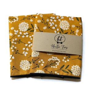 May include: Two mustard yellow fabric pocket squares with a white floral pattern and black trim. The pocket squares are folded and stacked on top of each other. A brown paper tag with the text "HoBo Ties" is attached to the top pocket square.