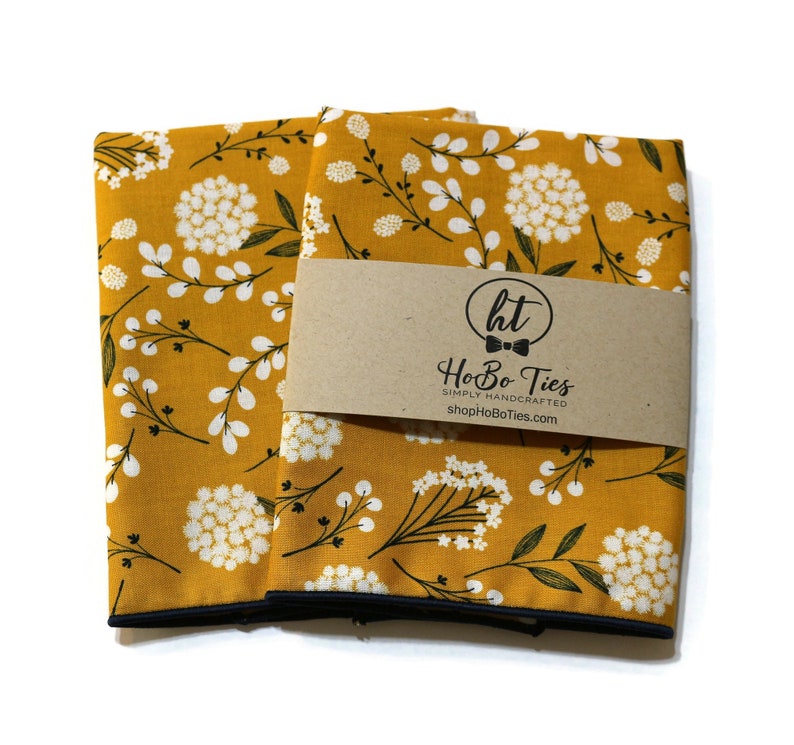 May include: Two mustard yellow fabric pocket squares with a white floral pattern and black trim. The pocket squares are folded and stacked on top of each other. A brown paper tag with the text "HoBo Ties" is attached to the top pocket square.
