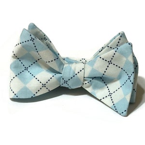 May include: A light blue and white argyle patterned bow tie. The bow tie is made of fabric and features a repeating diamond pattern with black dotted lines. The bow tie is tied in the center and is set against a white background.